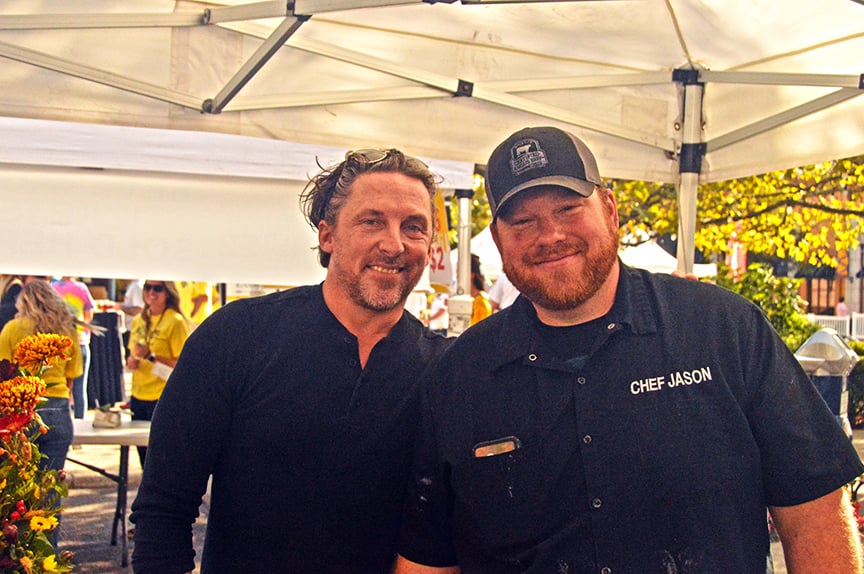 Two male chefs in black shirts, one with cap under white tent roofing at event