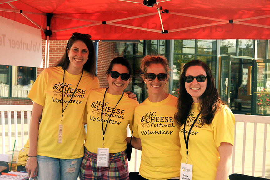 Four women under red tent roof wearing yellow mac and cheese fest t-shirts